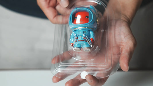 A person holding a clear blister package containing a blue astronaut figure with a red face.